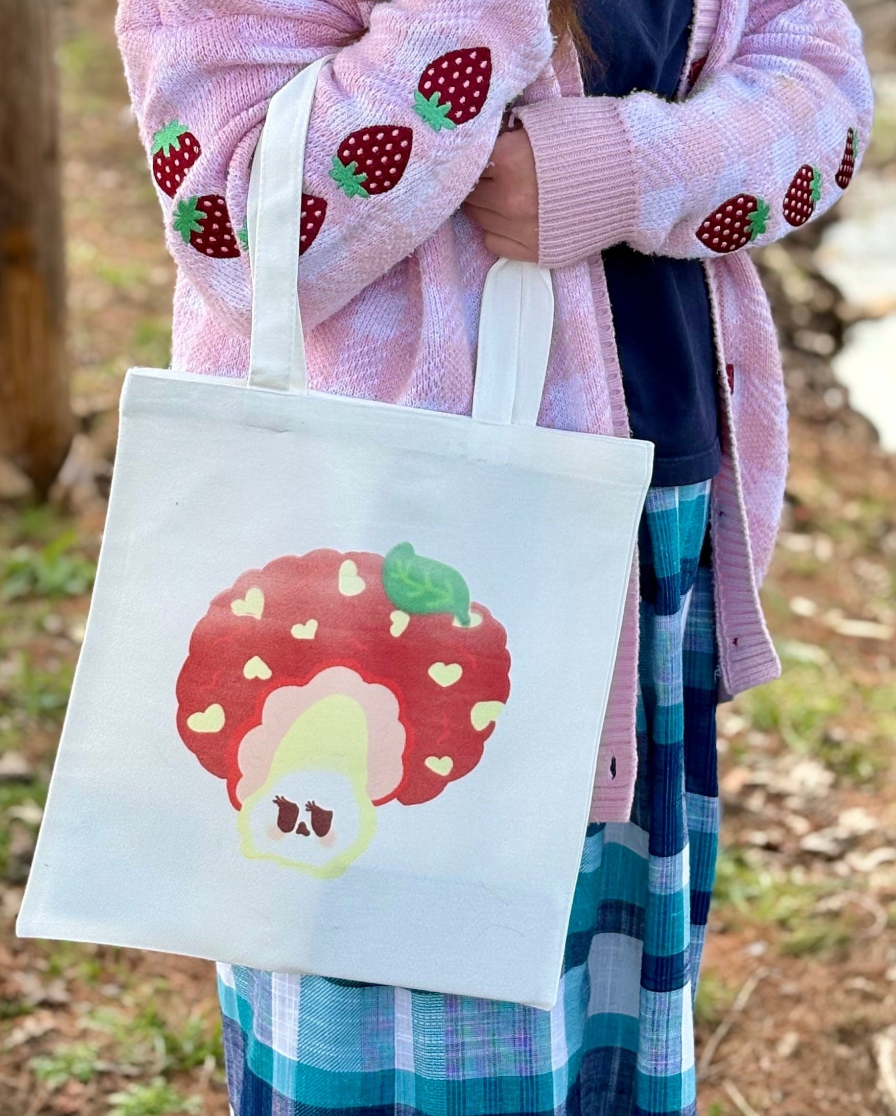 Person holding a tote bag with a strawberry design in a forest setting